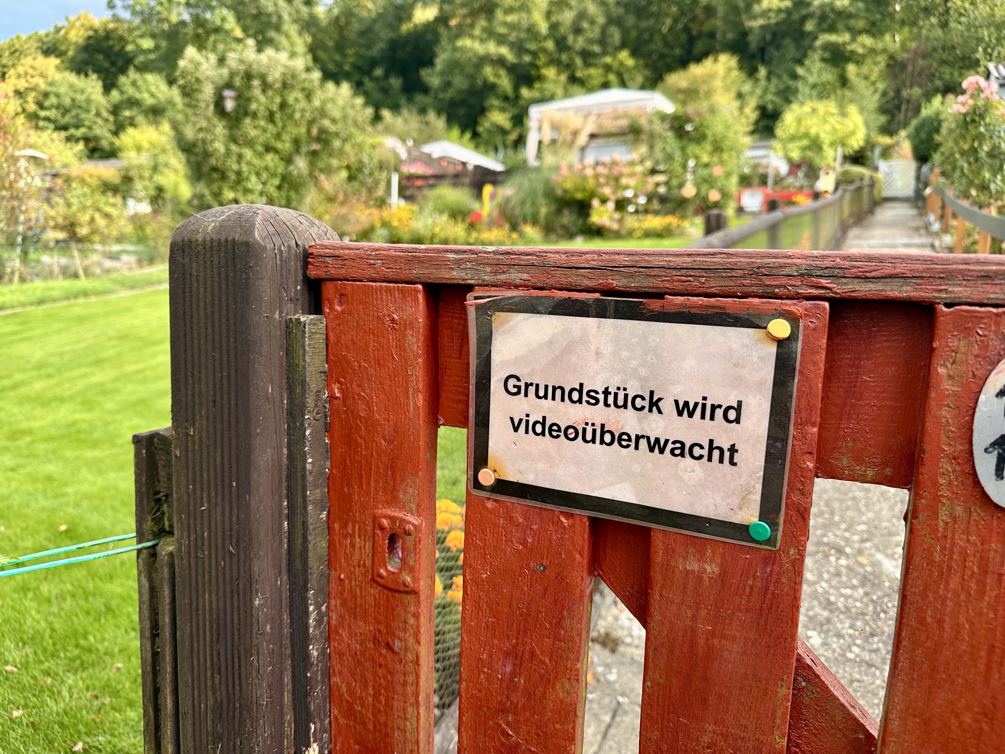 Videoüberwachung im Kleingarten – Hinweisschild am Eingangstor als Schutz vor Einbrüchen Schild mit der Aufschrift „Grundstück wird videoüberwacht“ an einem roten Gartentor im Kleingartenverein