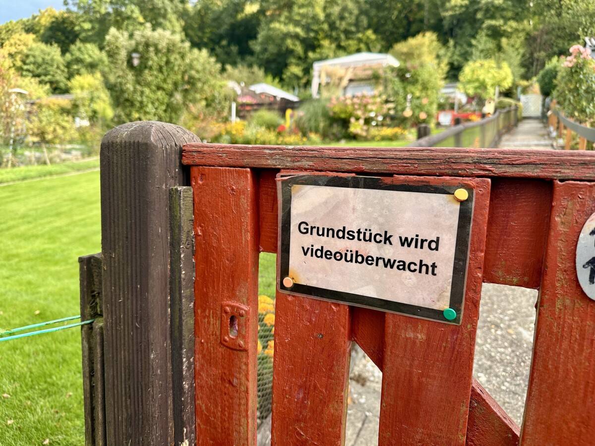 Videoüberwachung im Kleingarten – Hinweisschild am Eingangstor als Schutz vor Einbrüchen Schild mit der Aufschrift „Grundstück wird videoüberwacht“ an einem roten Gartentor im Kleingartenverein