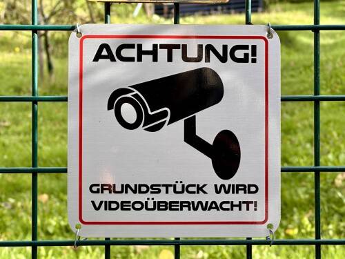 Warnschild mit Kamera-Symbol und Aufschrift „Achtung! Grundstück wird videoüberwacht!“ am Zaun